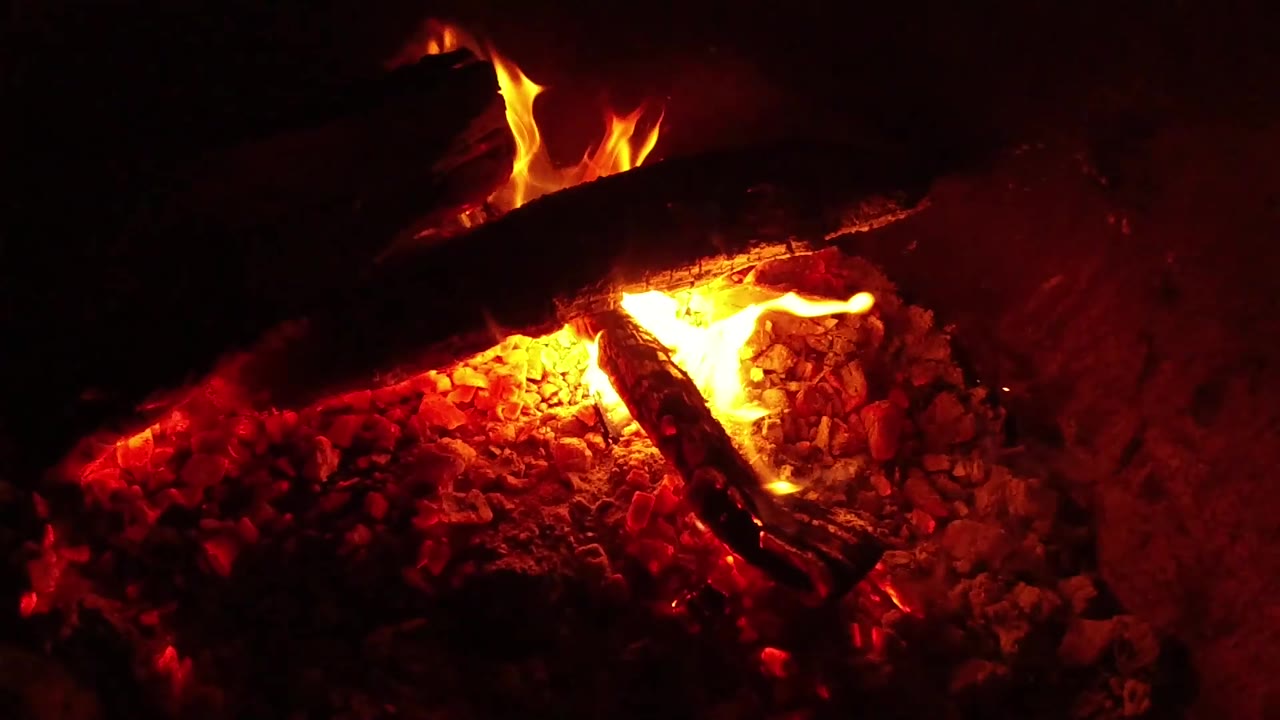 At 0:02, yellow-orange firelight flares along the logs, with the ember bed spread across the lower frame in Ojai, California, and no visible people.
