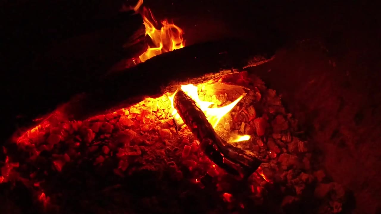 A taller orange flame lifts behind the upper charred log while red coals glow beneath the crossed wood in Ojai, California, with no visible people.