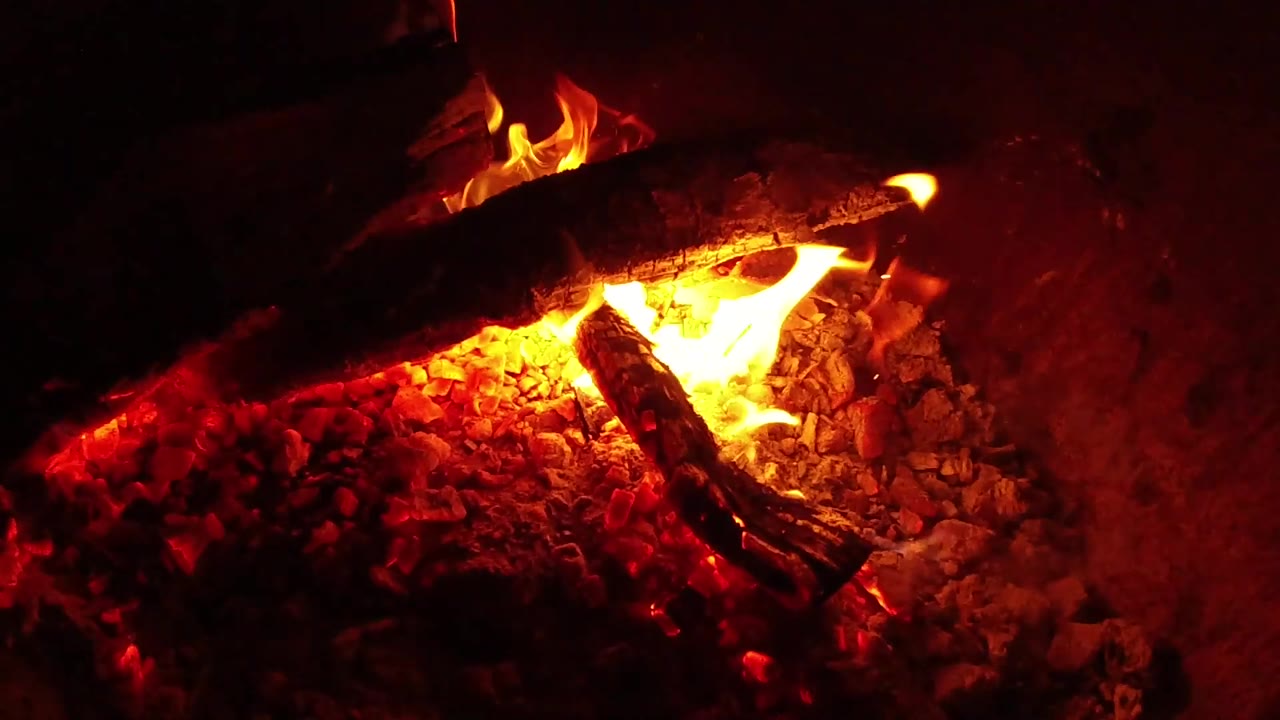 At 0:02, a bright yellow-orange flame curls under the top log and lights the surrounding coals in the Ojai, California fire pit, with no visible people.