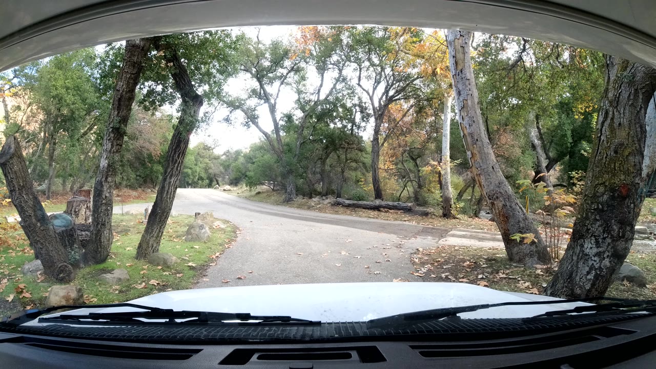 Ojai, California windshield view from inside a car, with the hood and wipers at the bottom, a pale road bending through trees and rocks, and no visible people.