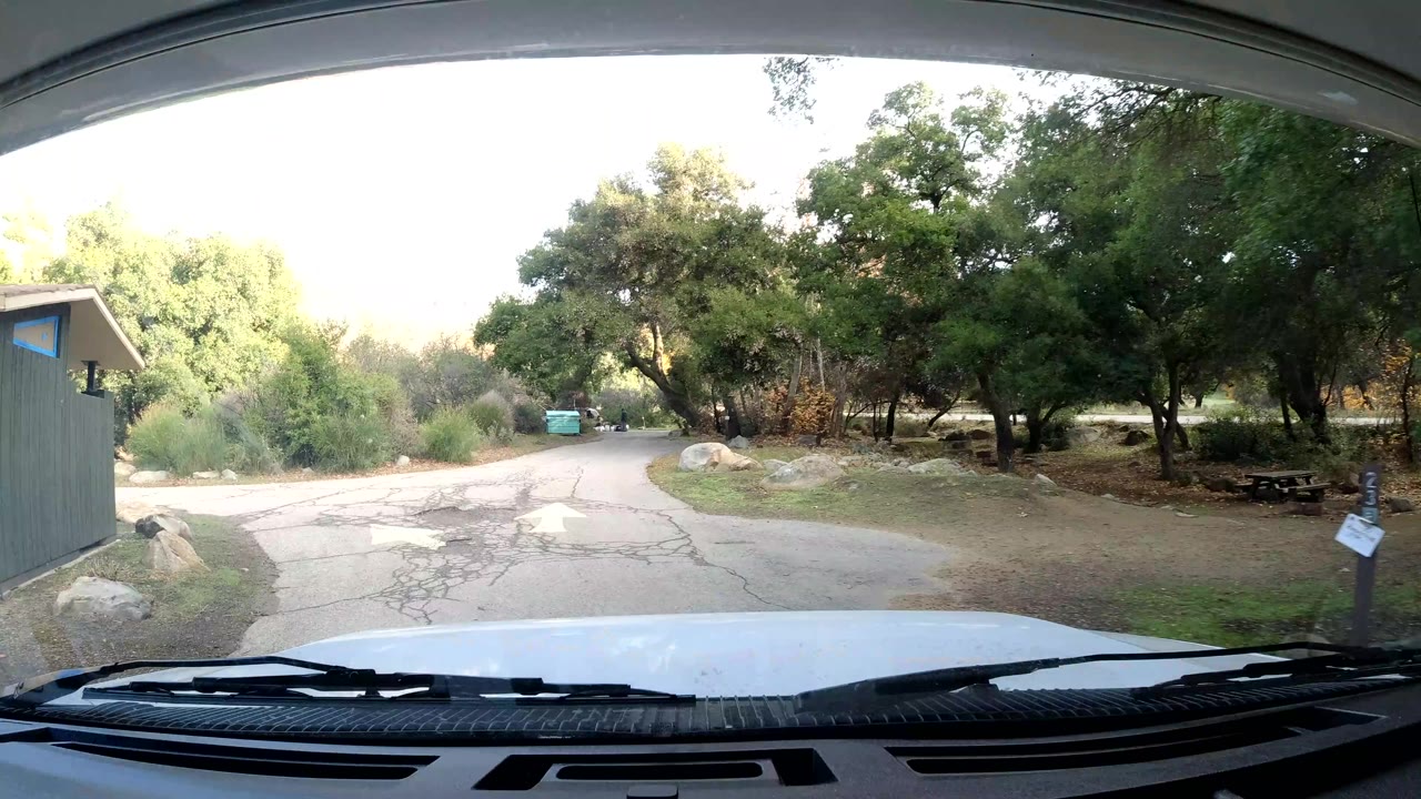 Ojai, California windshield view with a small roadside structure, cracked pavement, trees, boulders, signs, and no visible people.