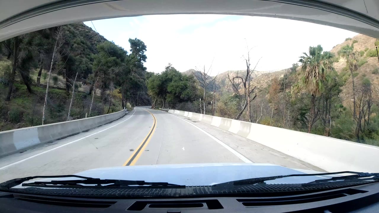 Vehicle POV of a right-curving Ojai road bordered by a concrete barrier, hillside trees, dry brush, and canyon slopes, with no visible people.