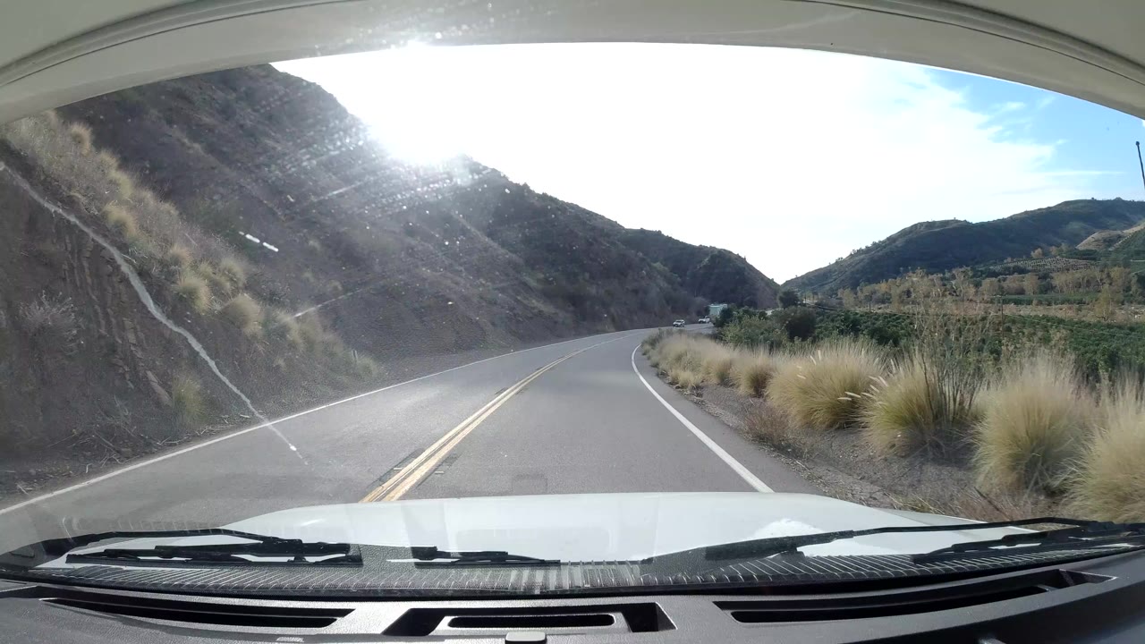Vehicle-mounted view through a windshield on a curving Ojai, California two-lane road with dry hillside, roadside grasses, bright sun glare, and no visible people.
