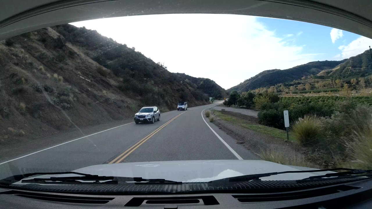 Vehicle POV on an Ojai, California road with oncoming cars, a dry hillside on the left, planted grasses on the shoulder, and no visible people.
