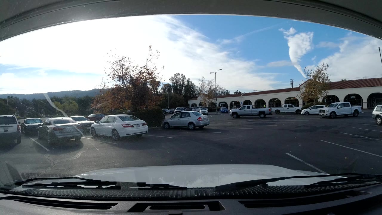 Opening view of an Ojai, California shopping center parking lot through a windshield, with parked cars, arched storefronts, bright sun glare, distant hills, and no visible people.