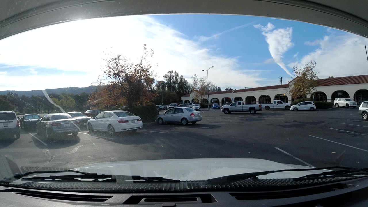 Wide Ojai shopping center parking lot frame with a white vehicle at right, cars across the asphalt, arched storefronts, trees, hills, strong glare from the sky, and no visible people.