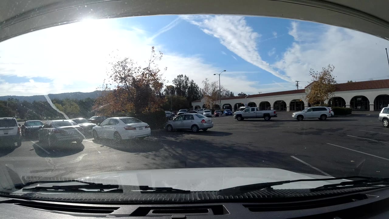 Parked vehicle windshield frames cars, trees, arched storefronts, and sun glare.