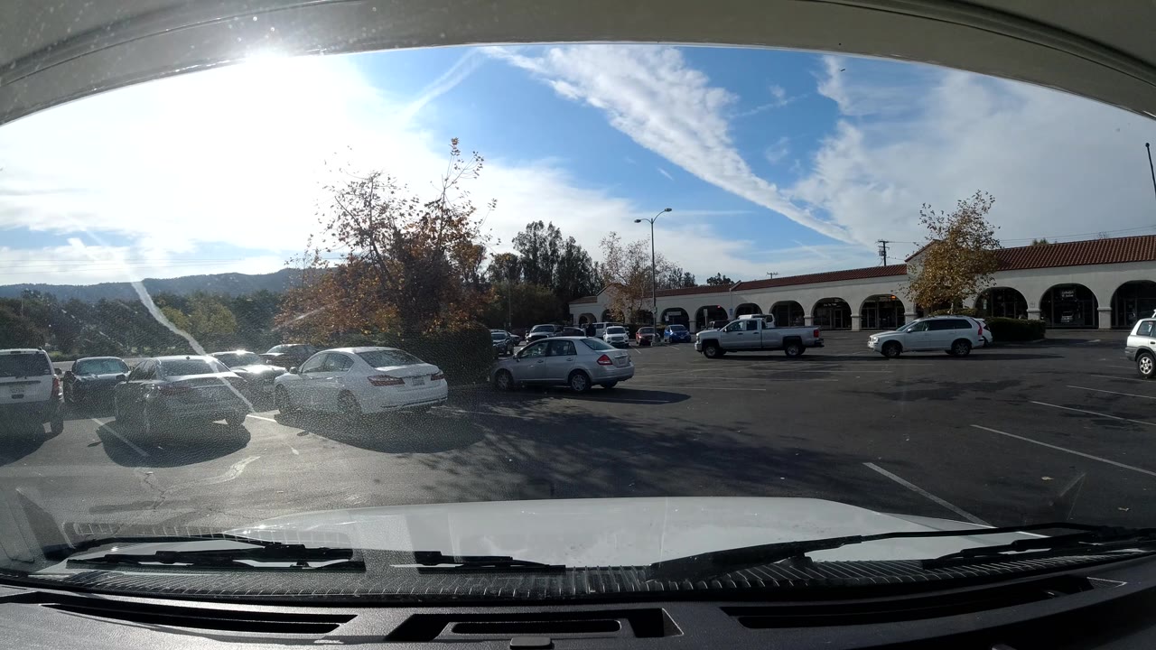 At 0:02, the Ojai, California lot remains framed by the dashboard and windshield, with bright glare on parked vehicles, a low arched building, hills, and no visible people.