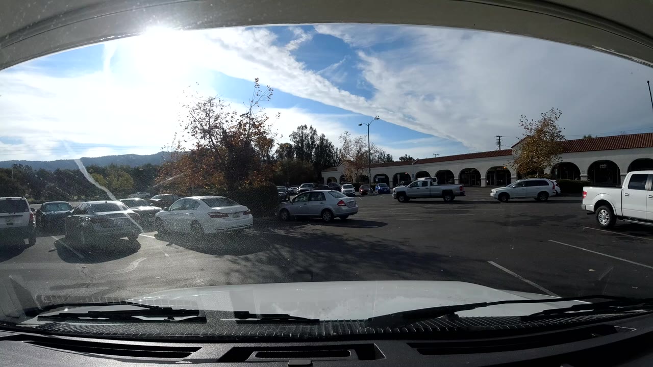 Opening frame of an Ojai, California parking lot seen through a vehicle windshield, with cars, arched storefronts, trees, hills, bright glare, and small distant people.
