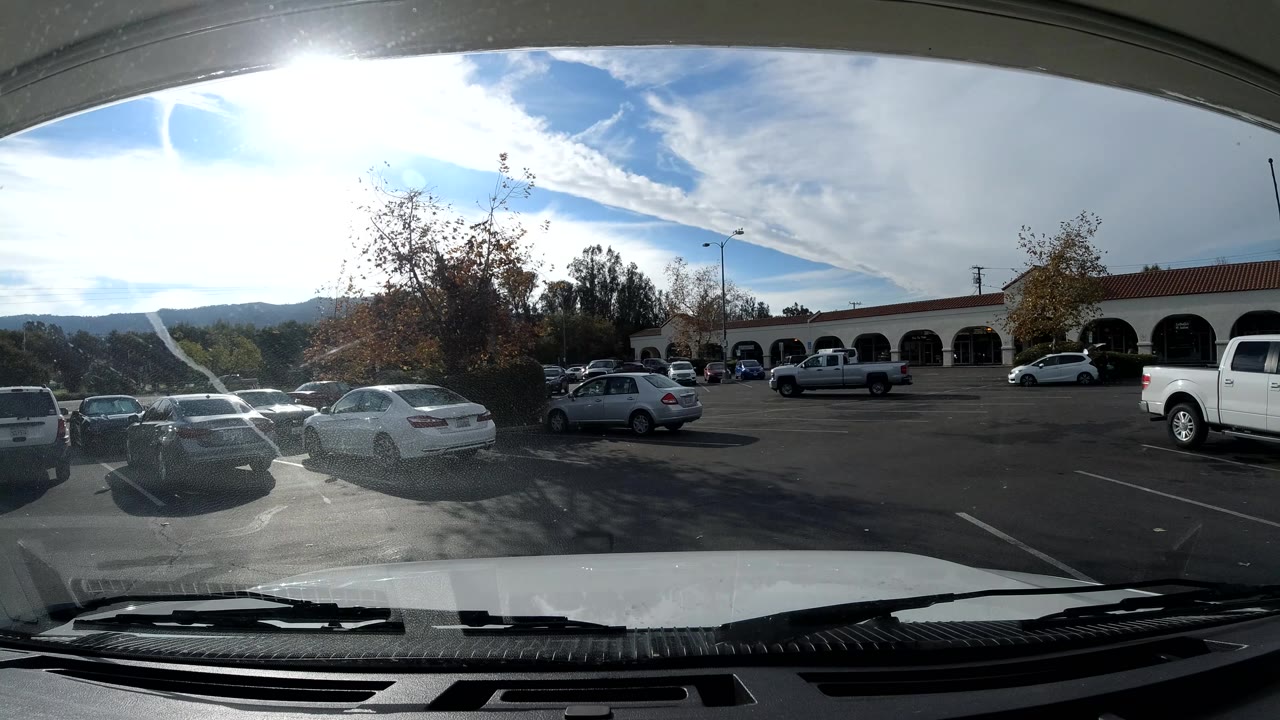 Third frame of the Ojai street-view time-lapse has stronger sun flare over the windshield, with cars in the lot, trees near the storefronts, low hills, and arched building fronts.