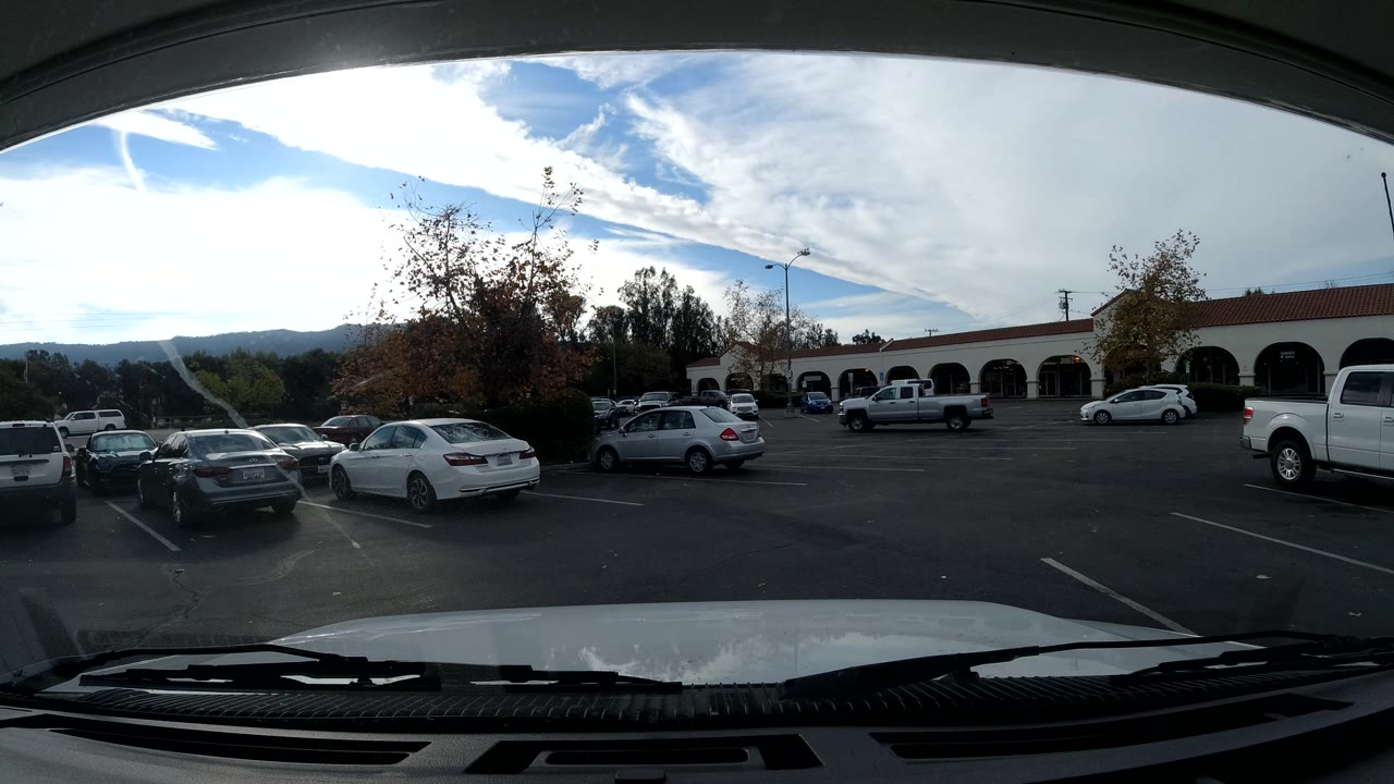 Parking lot with several cars, a white pickup, arched storefronts, trees, and distant hills.