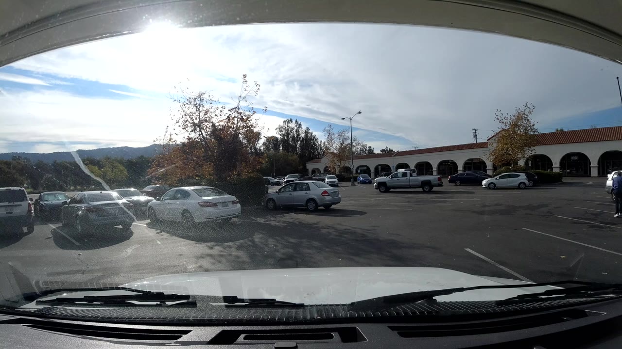 Ojai, California parking lot seen through a vehicle windshield, with a white hood and wipers in front, parked cars across the pavement, arched storefronts, trees, distant hills, and small people near the building.