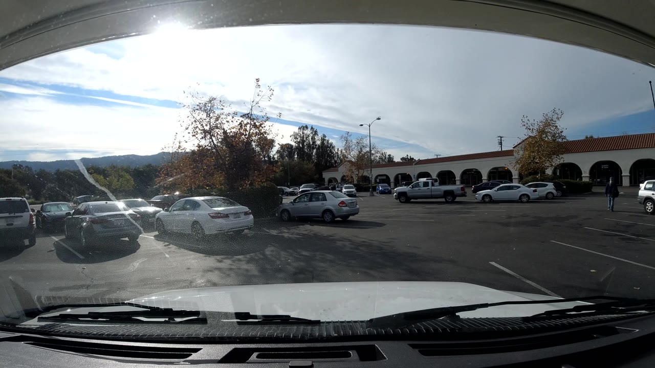 Ojai, California parking lot under bright clouds, with windshield glare, parked cars in rows, trees near the center, an arched building across the background, hills beyond it, and a small person near the right edge.