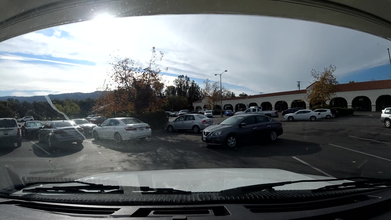Parking lot through a windshield with a dark car closer in the foreground and arched storefronts behind.