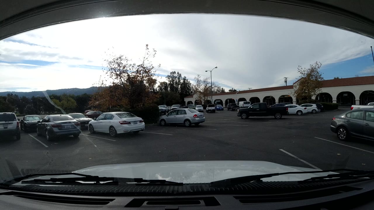Front-mounted vehicle view in Ojai, California showing a parking lot with several cars, arched storefronts, distant hills, and no clearly visible people.
