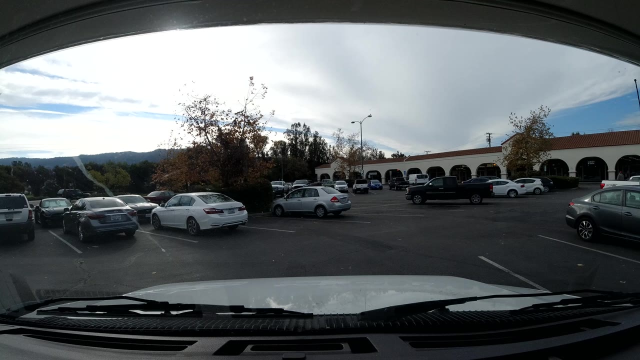 Vehicle-mounted view in Ojai, California continuing across a parking lot with parked cars, low storefront arches, trees, hills, and no clearly visible people.