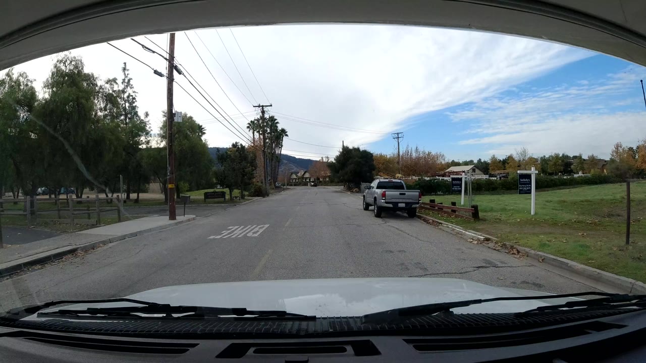 Driver's-view frame in Ojai, California showing a straight street, parked trucks, utility poles, trees, a grass area, and no visible people.