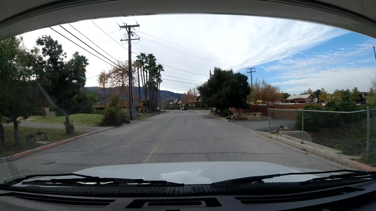 Forward windshield view in Ojai, California with utility poles, trees, low roadside buildings, asphalt pavement, and no visible people.