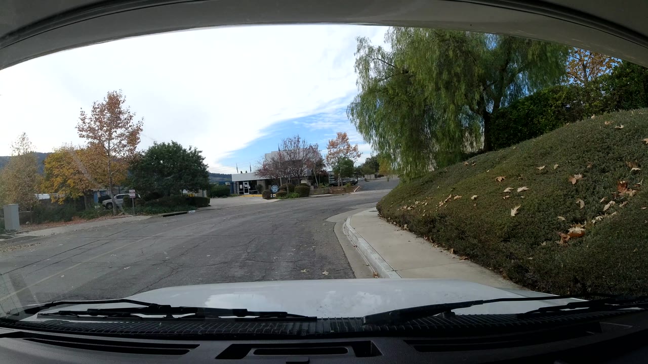 Windshield view in Ojai, California as the street curves near a chain-link fence, trees, a hedge, parked vehicles, and no visible people.