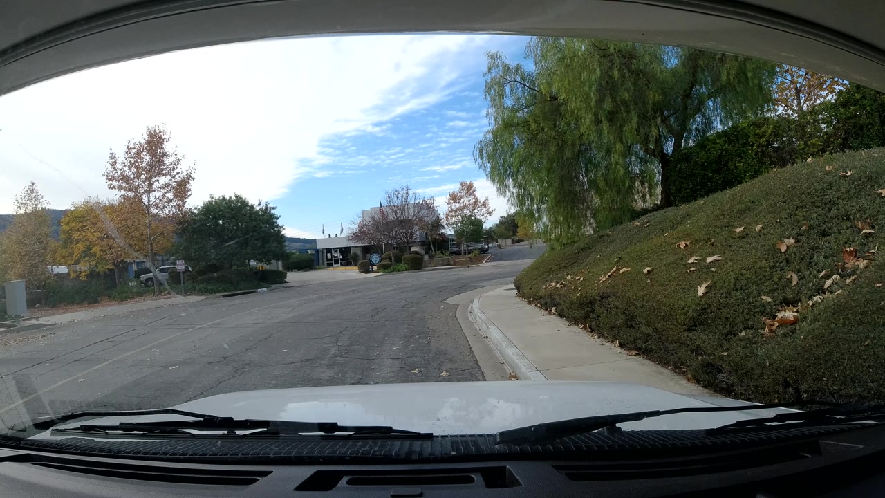 Inside-car view of an empty Ojai, California road with sidewalk, hedges, autumn-colored trees, distant hills, and no visible people.