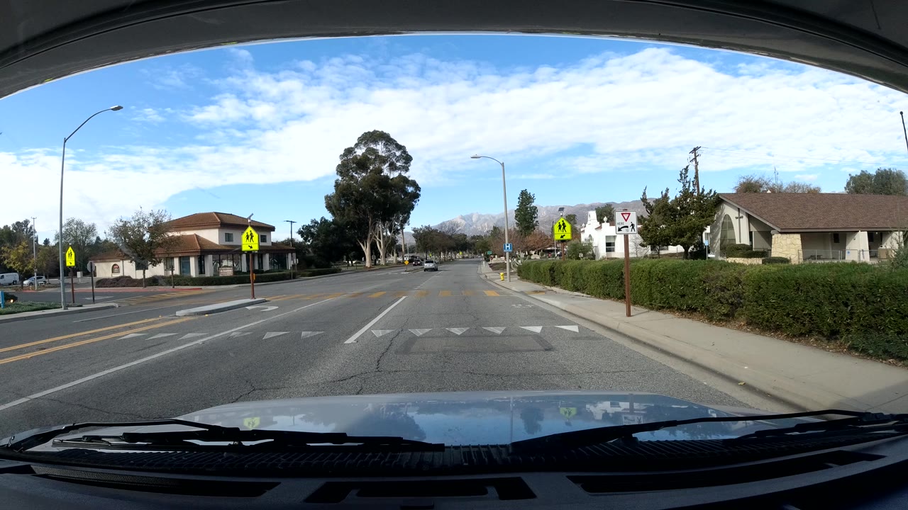 A windshield view in Ojai, California shows a two-lane road with crosswalk markings, pedestrian signs, roadside homes, trees, and no visible people.