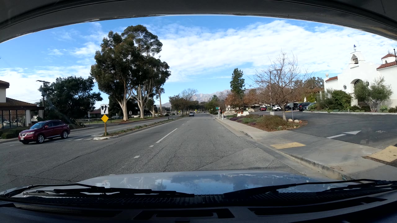 Vehicle approaches a tree-lined road with a white building at right.