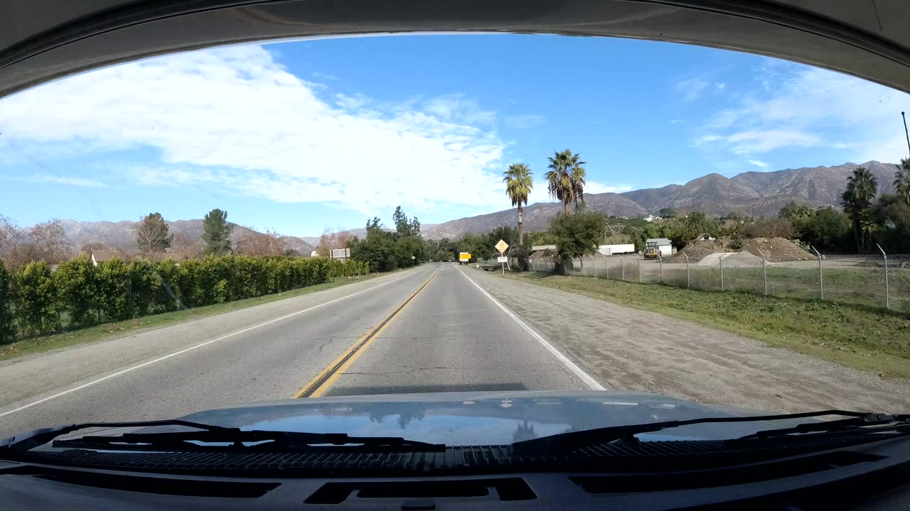 A windshield view in Ojai, California looks down a two-lane road with a hedge line, palms, open roadside ground, distant hills, and no visible people.