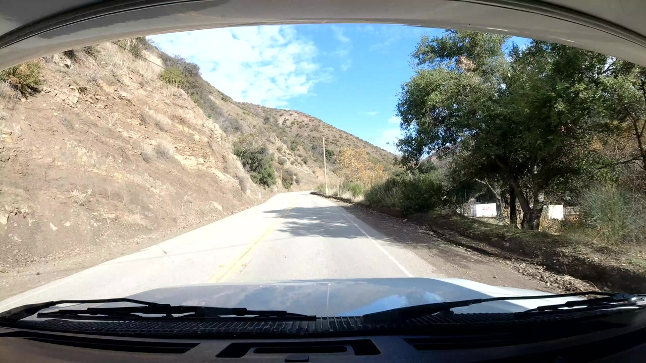 Sunlit vehicle POV on an Ojai mountain road with a pale exposed hillside to the left, trees to the right, and no visible people.