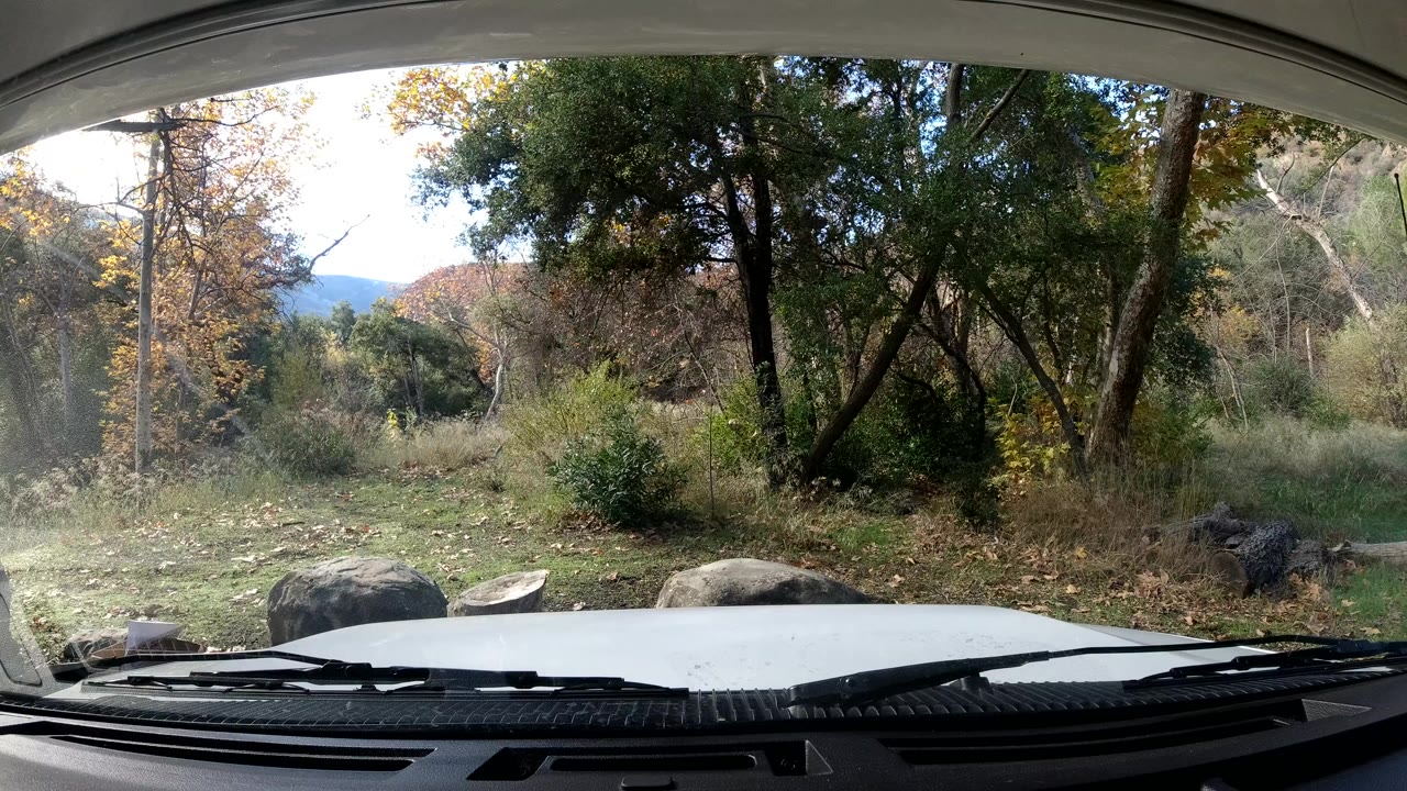 At one second, the Ojai windshield view holds on dense green foliage, pale grass, foreground rocks, and the car hood, with no visible people.