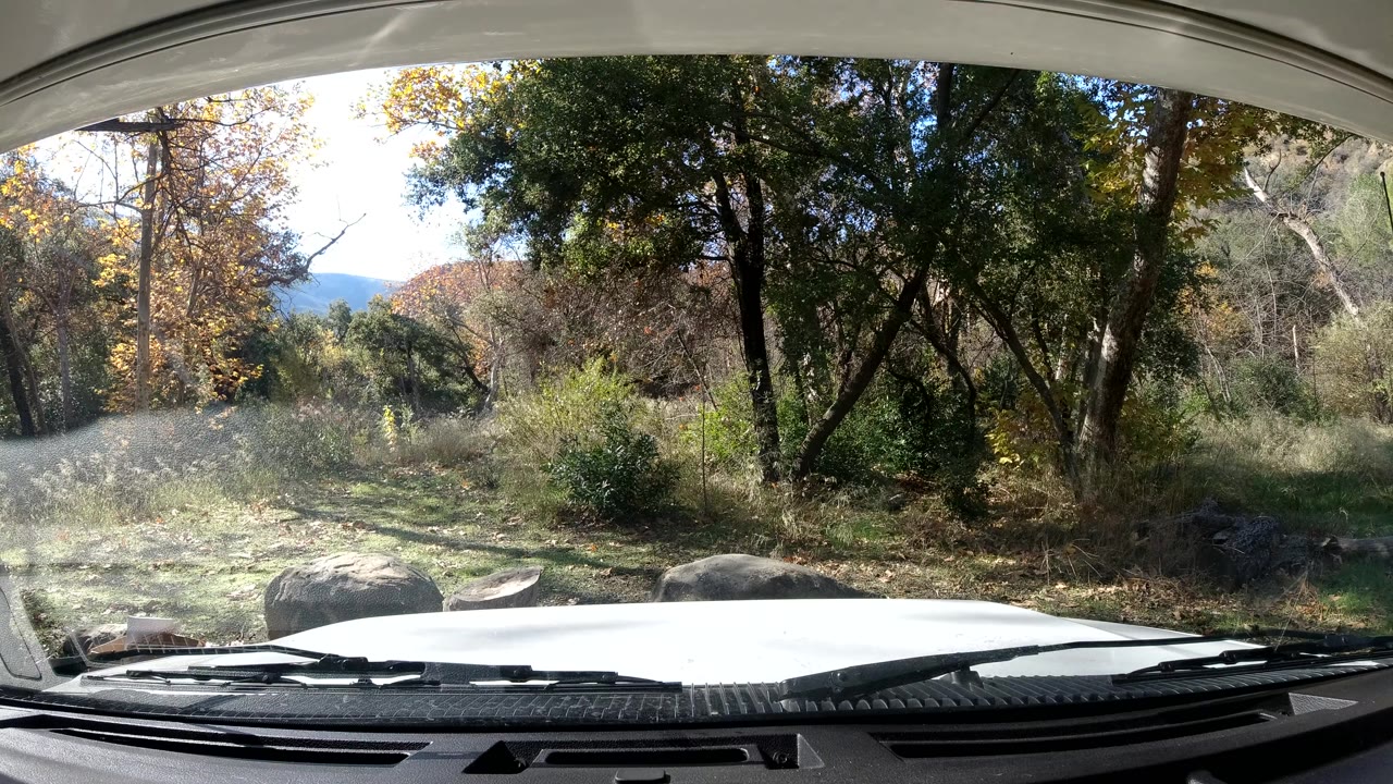 Third Ojai, California frame showing the dashboard and white hood below a windshield view of trees, dry grasses, boulders, and no visible people.