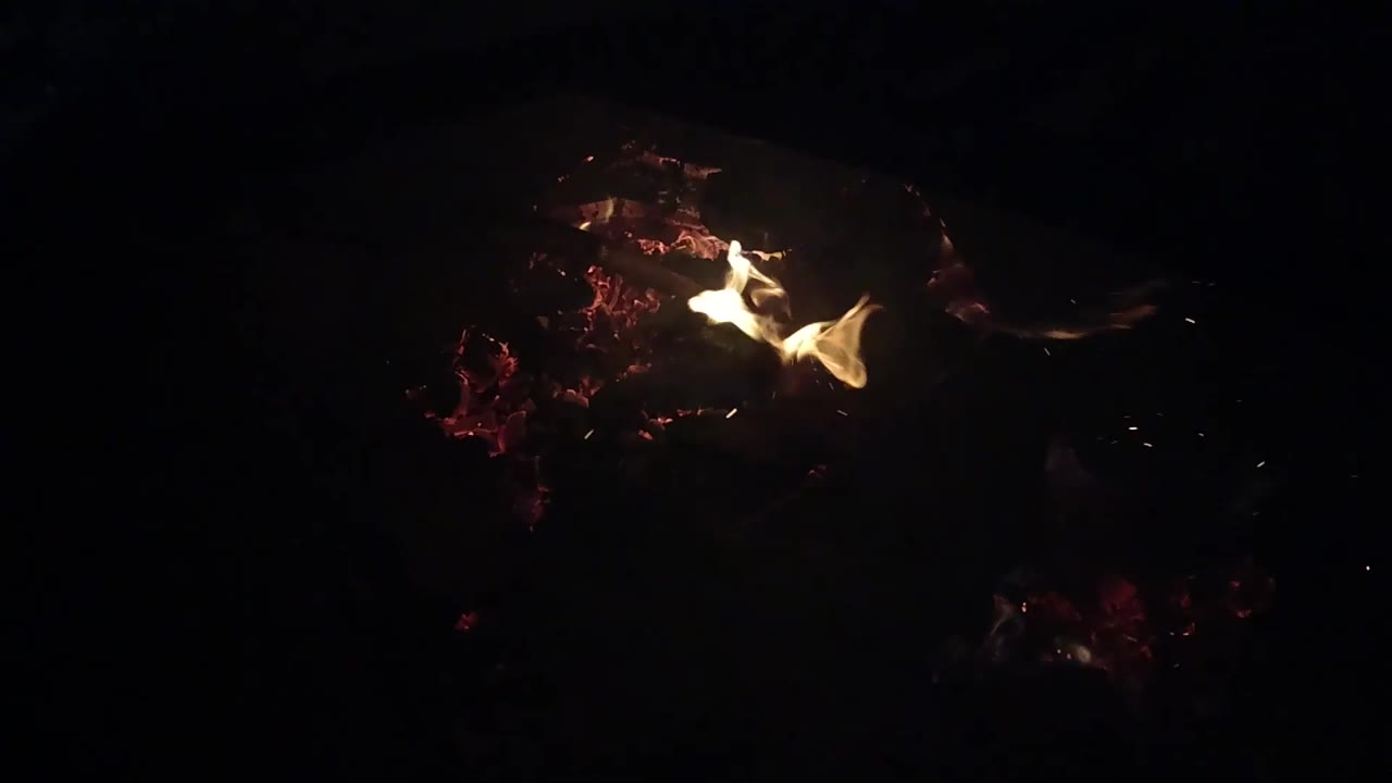 A smaller flame curls above dim red embers in the dark Ojai fire frame, leaving much of the image black and no visible people.