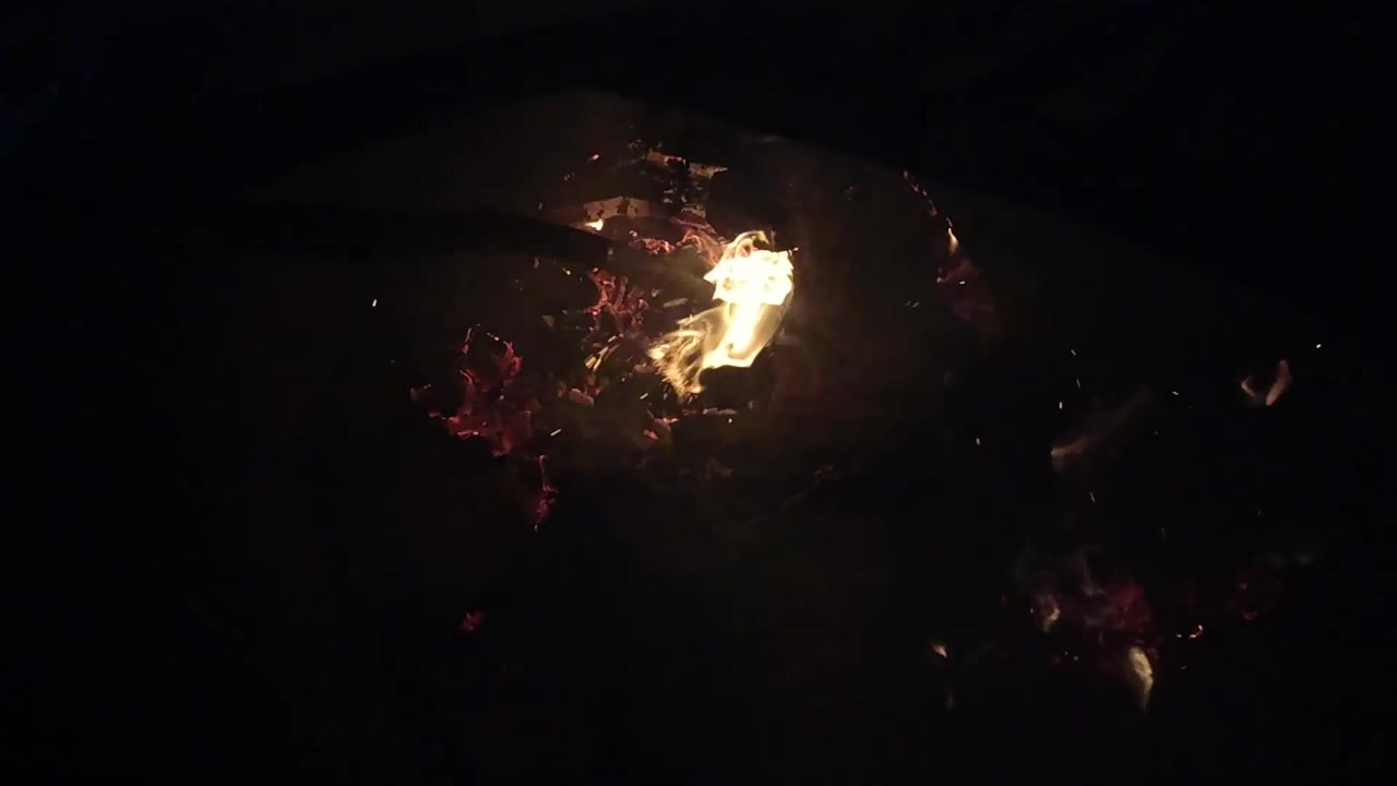 A flame rises from a compact cluster of red embers in the Ojai night-fire view, with the rest of the frame falling dark and no visible people.