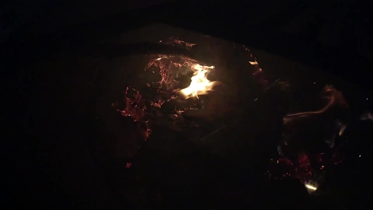 A brighter flame curls sideways across charred material and red embers in the Ojai night campfire, with no visible people.