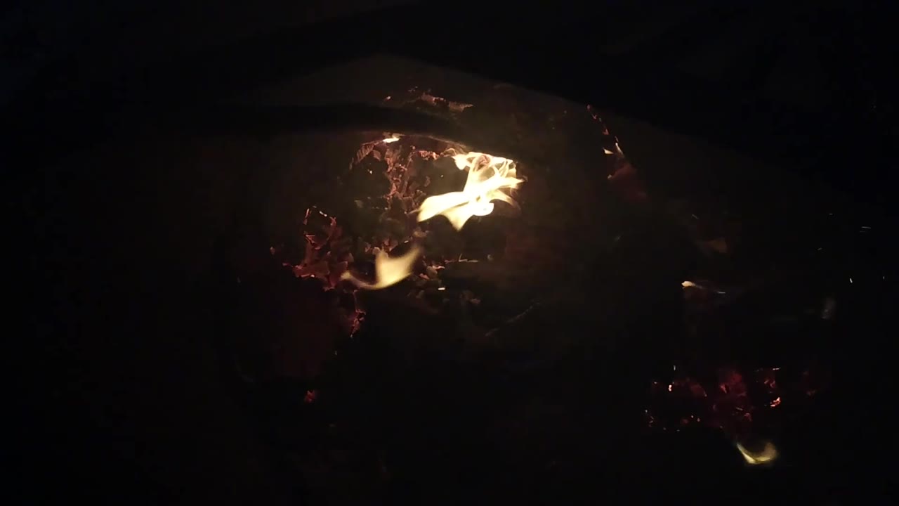 A flame arcs above the ember cluster in a mostly black Ojai, California campfire close-up, with no visible people.