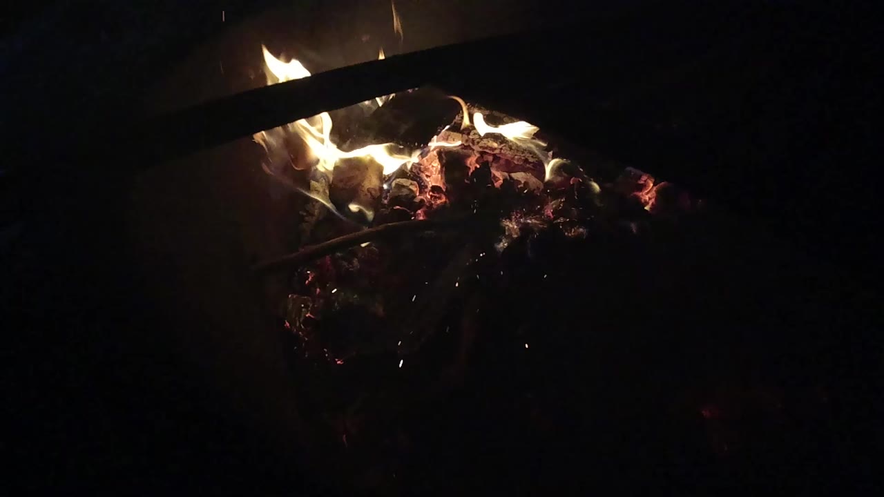 At 0:00, small yellow flames burn over red embers in Ojai, California, with a dark diagonal object crossing the upper frame and no clearly visible people.