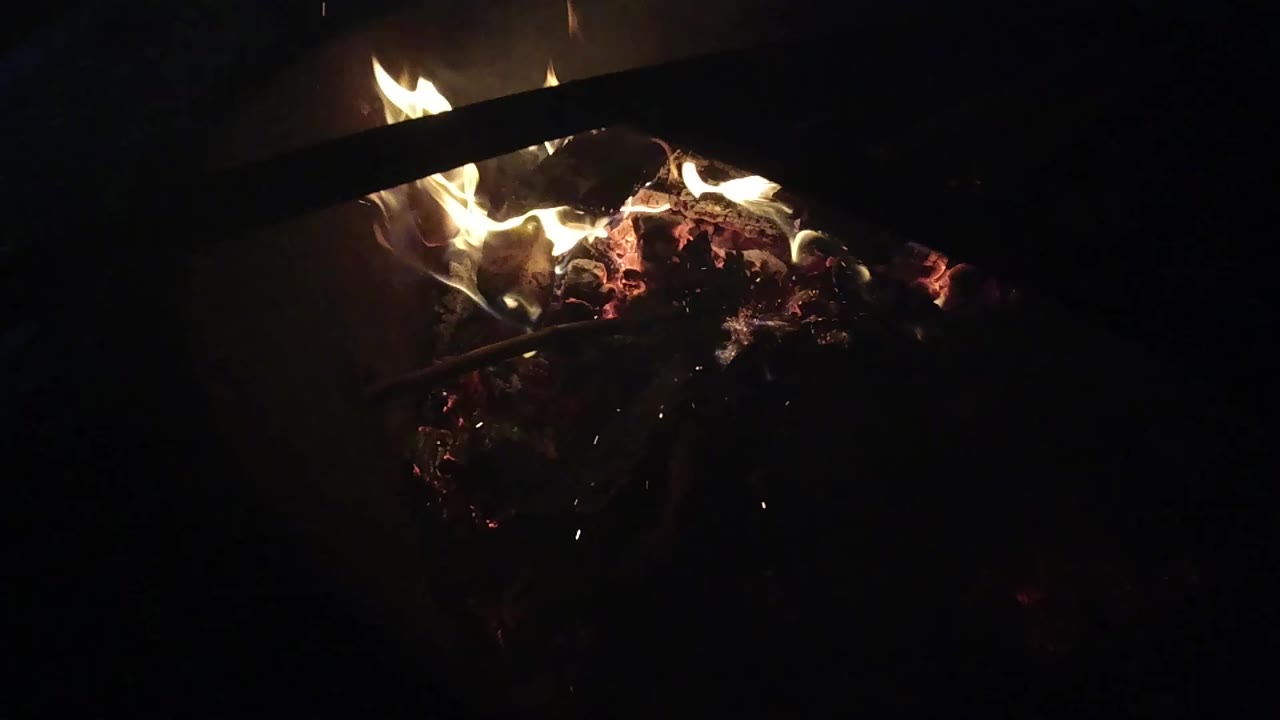 At 0:01, the Ojai fire remains tight in frame, with pale flames licking over a red ember bed and surrounding detail falling into deep shadow.