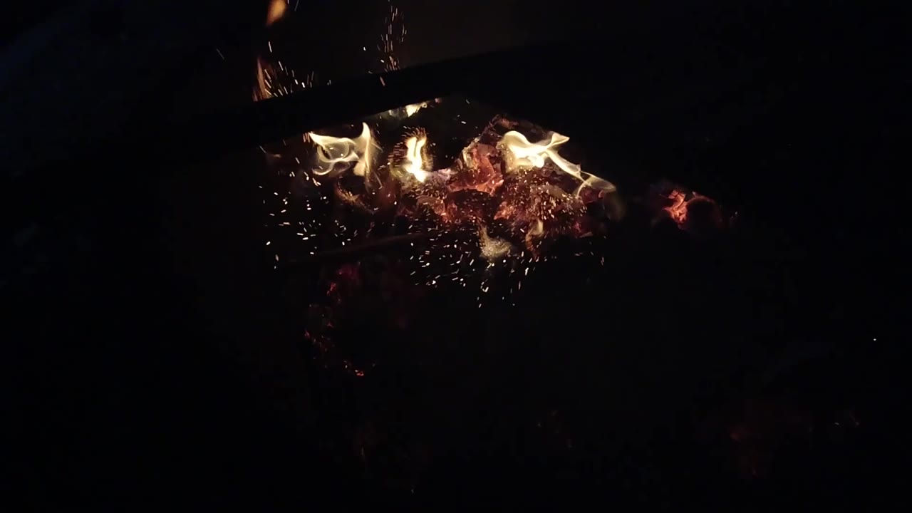 Sparks rise beside a small night fire and ember bed.