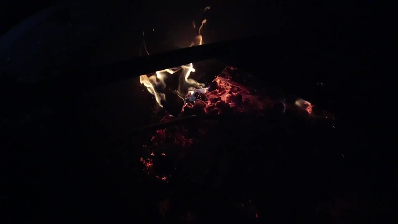 At 0:03, several flame tongues flare over the red ember bed in Ojai, California, beneath a dark diagonal log or bar and with no visible people.