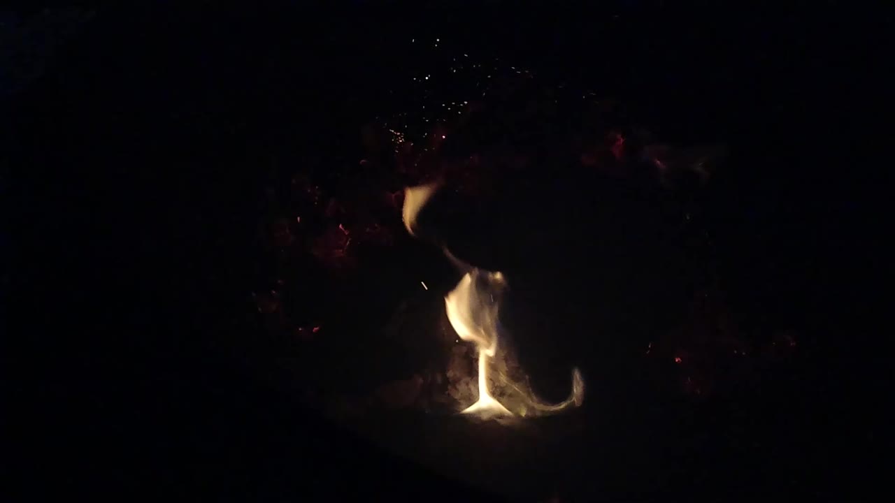 The flame brightens at the base and leans upward in the dark Ojai fire pit, with scattered ember points and no visible people.