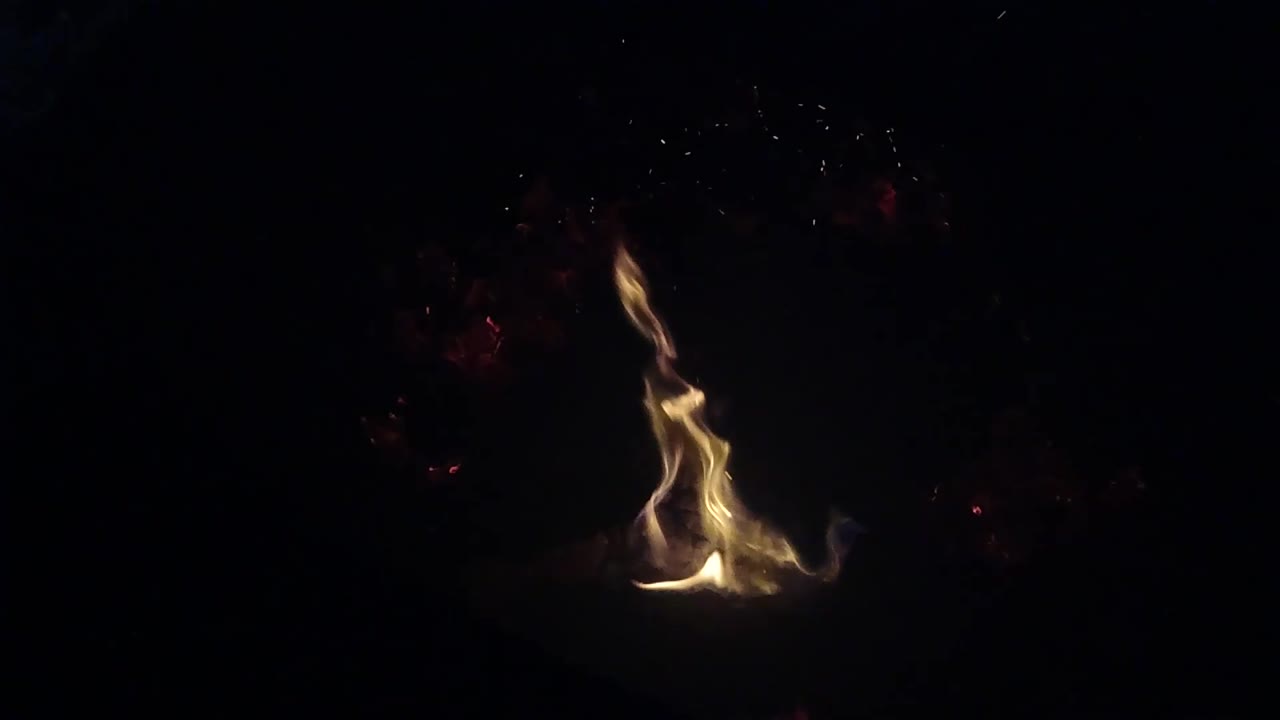 A taller narrow flame rises from the ember bed in Ojai, California, with a few dim red coals visible nearby and no visible people.
