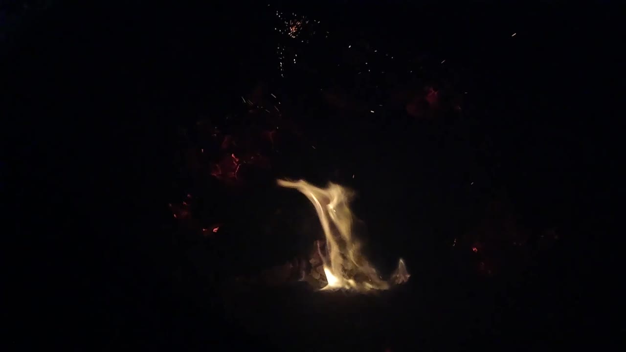 The flame bends left across a low ember bed in the dark Ojai night frame, with red coals glowing faintly and no visible people.