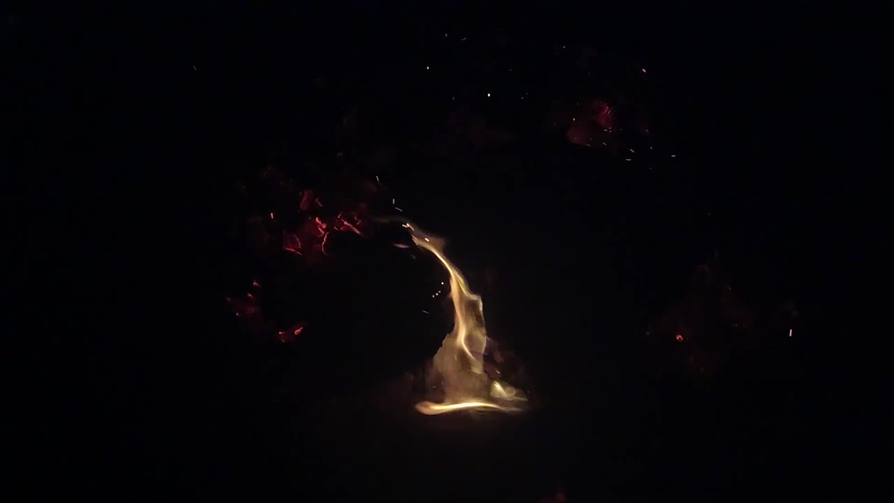 A taller flame curls to the right above a small bright fire in the Ojai, California night scene, with scattered red embers and no visible people.