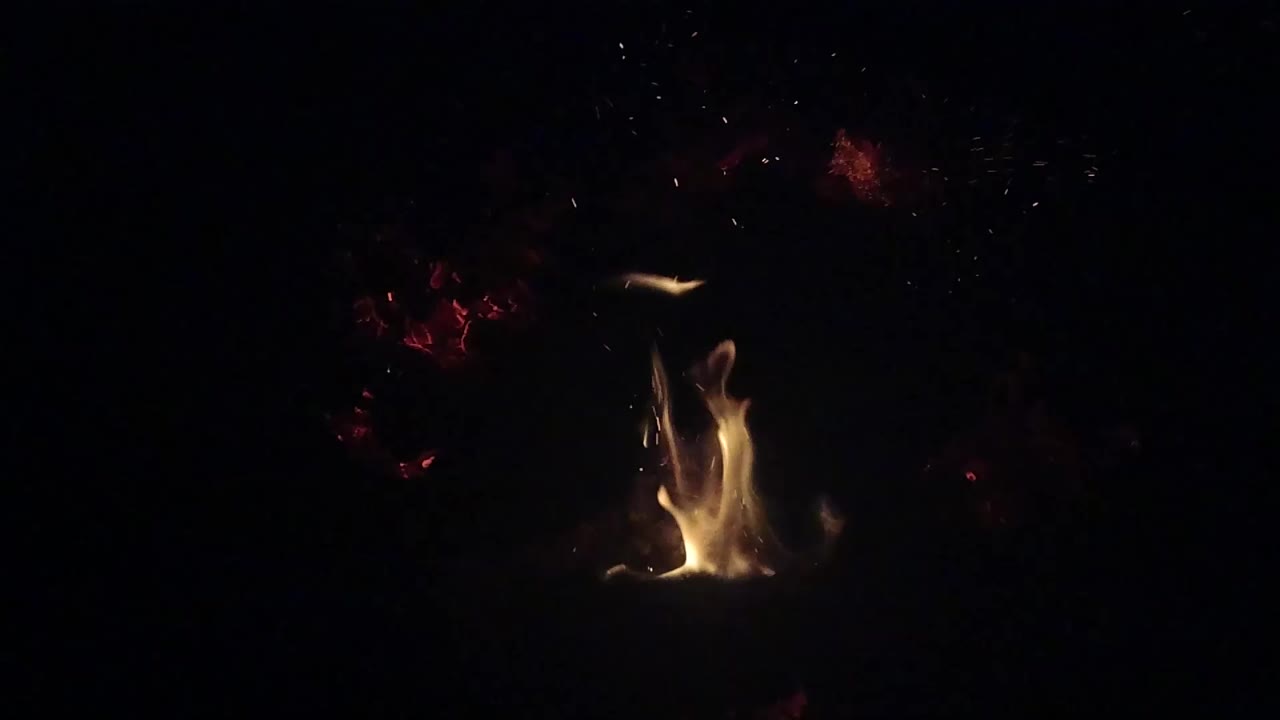 A yellow campfire flame rises from a small fire in Ojai, California, with red embers scattered in the dark background and no visible people.