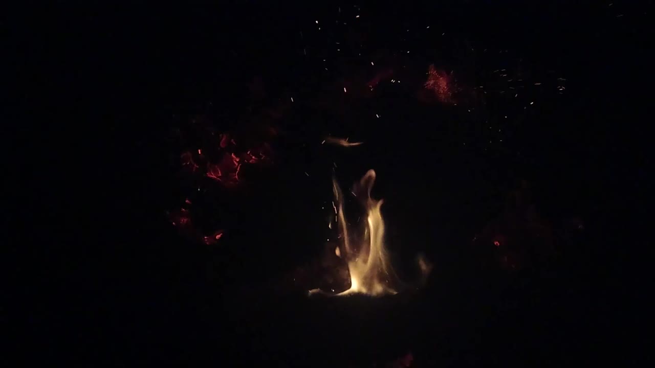 A taller yellow flame curls upward from the Ojai campfire, with faint red embers behind it and no visible people.