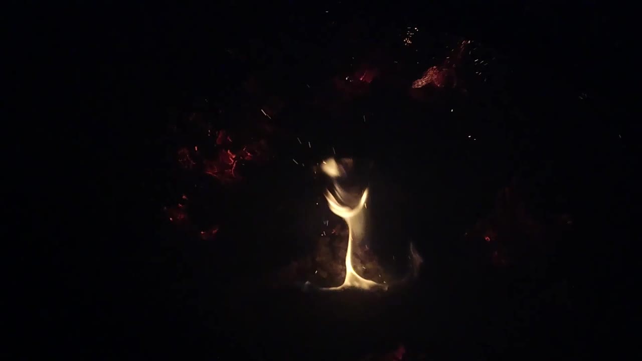 A narrow yellow flame leans to the right over the small Ojai campfire, with sparse sparks and red ember patches visible in the darkness and no visible people.