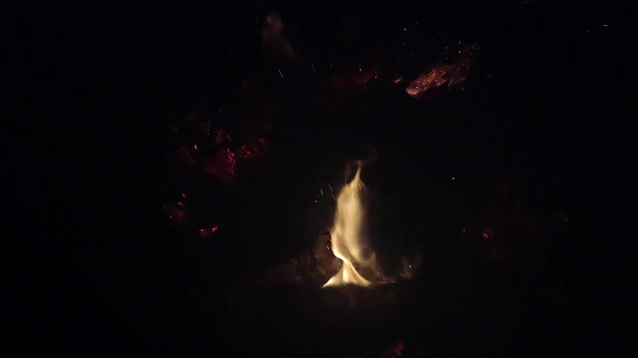 A compact yellow flame rises from the low Ojai campfire, with dim red embers above and around it and no visible people.