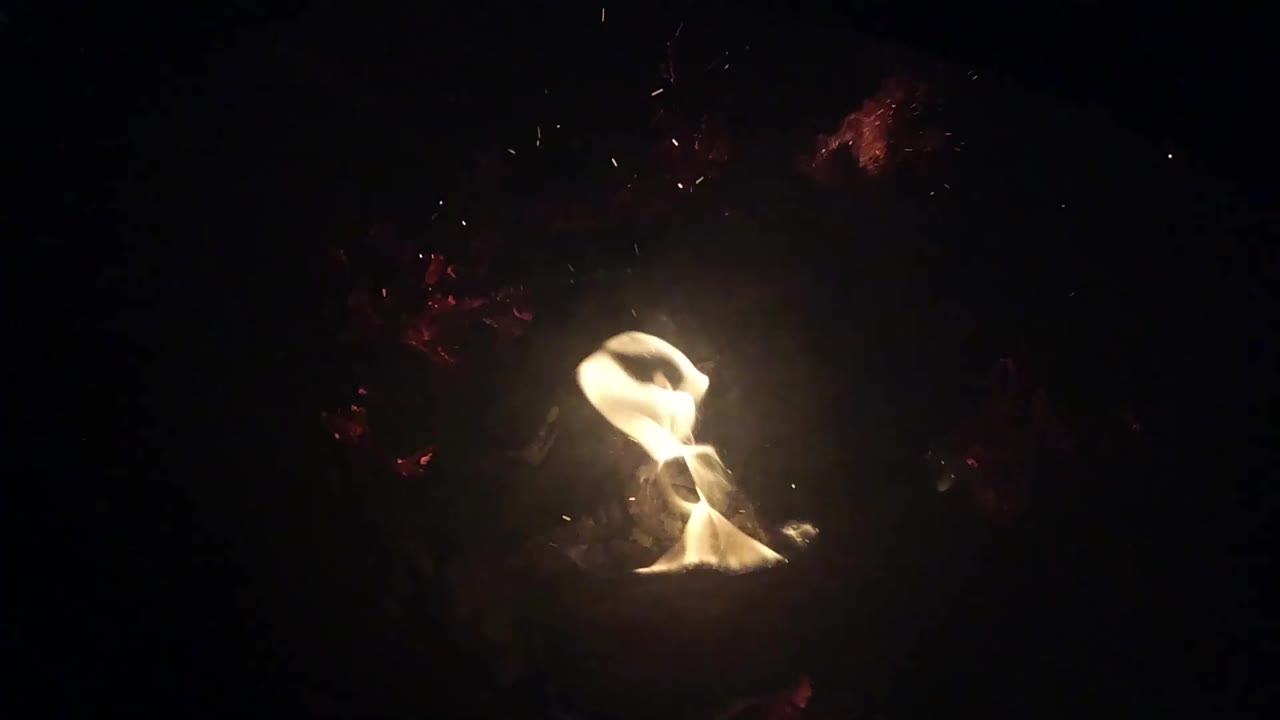 A brighter flame rises from charred branches and leaf litter in Ojai, California, surrounded by darkness and no visible people.