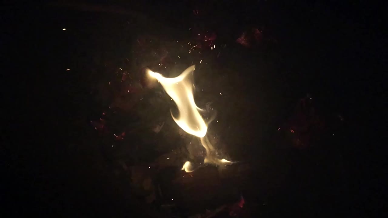 A curled yellow flame glows above ash and charred material in Ojai, California, surrounded by deep darkness with no visible people.