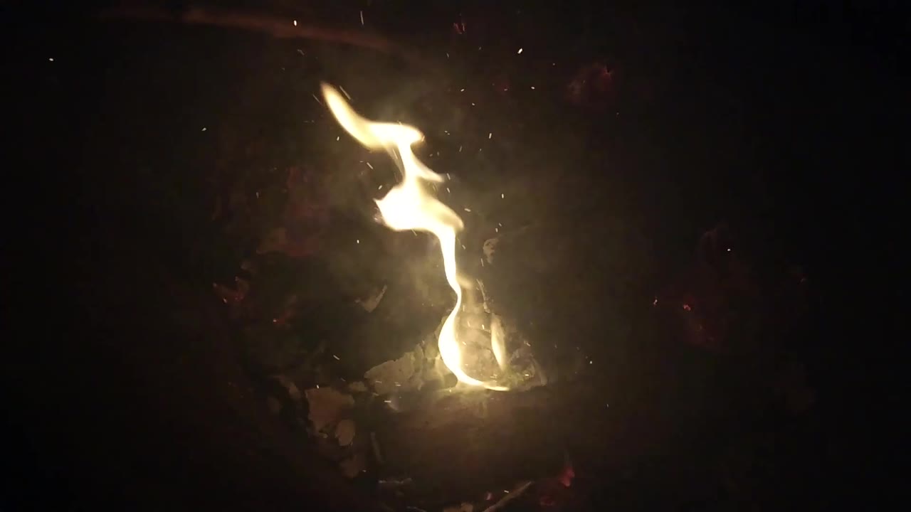 A taller yellow flame lifts from a small glowing fire bed in Ojai, California, with dark surrounding ground and no visible people.