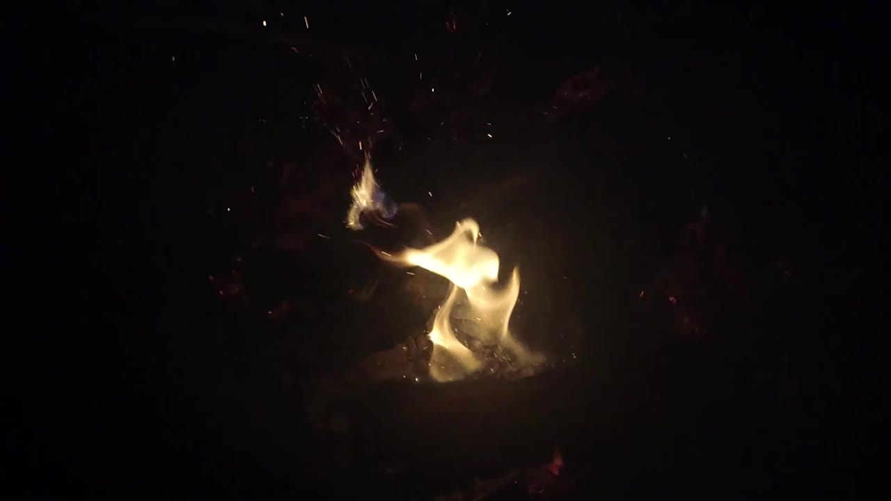 A low flame burns beside charred wood and pale ash in Ojai, California, with the background falling almost completely black and no visible people.